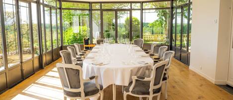 A large, round dining table set for six with white tablecloths and grey upholstered chairs, surrounded by large windows offering a view of greenery.