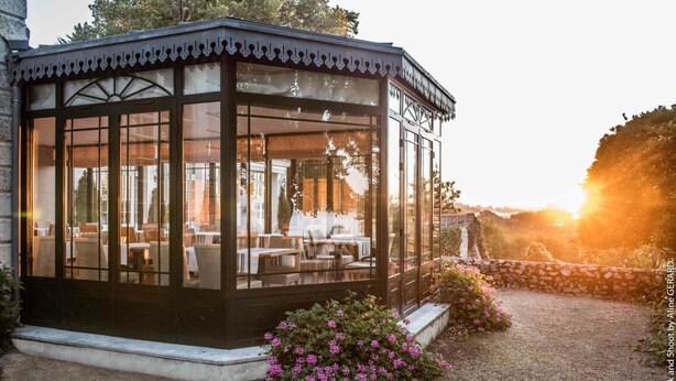 Un belvédère en verre, doté de tables et de chaises, entouré d’un mur de pierre et de plantes en fleurs.