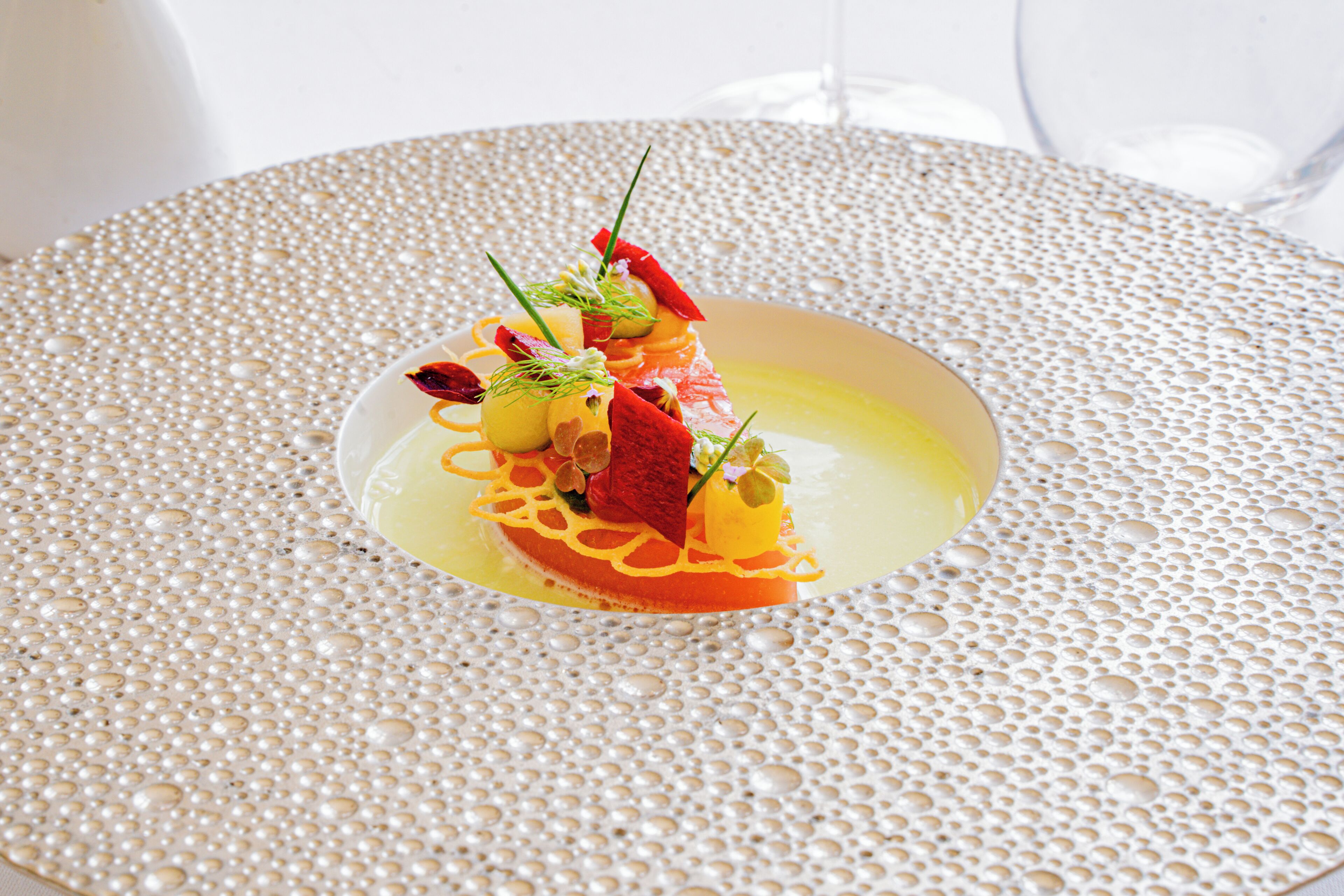 A plate of food with various garnishes and a clear liquid on a textured surface.