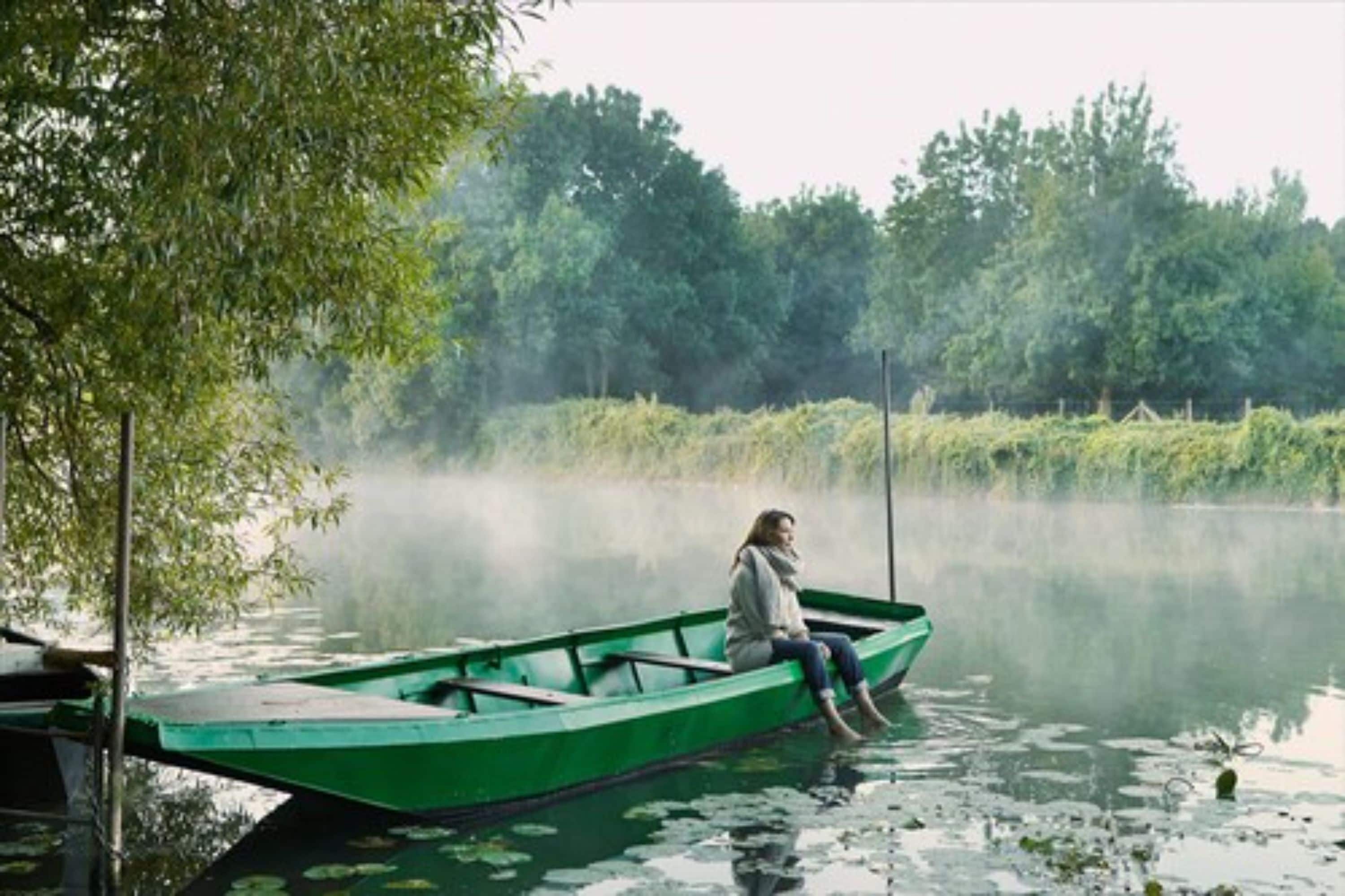 Iemand zit in een groene boot op een kalm meer, omringd door bomen.