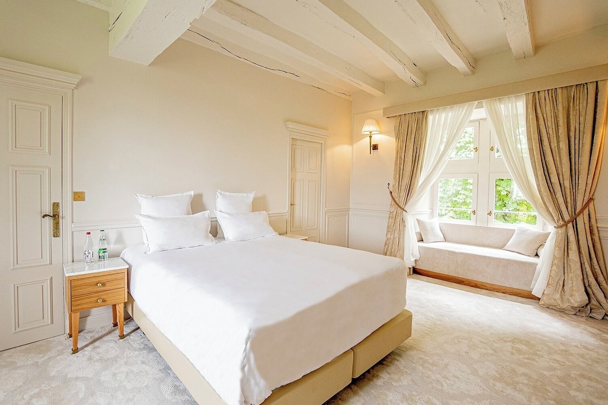 Ein Schlafzimmer mit einem großen Bett, einem Nachttisch, einem Fenster mit Vorhängen und einer fest eingebauten Sitzbank am Fenster.