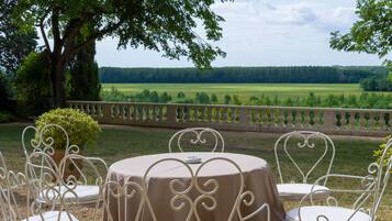 Mesa redonda con mantel beige y sillas de metal blanco, ubicada al aire libre con vistas a un campo verde y árboles al fondo.