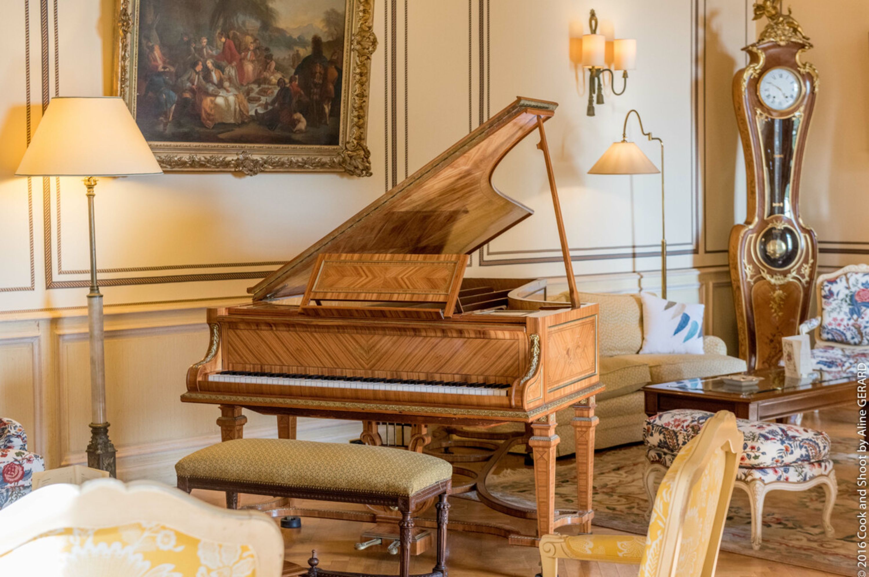 A grand piano in a well-appointed room with a sofa, armchairs, and a large clock.