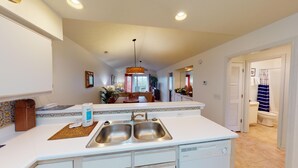 A kitchen with a double sink, white countertops, and a refrigerator. There is a dining area with a table and chairs visible through the kitchen. - Secluded Paradise Serene 2BR Golf Course Views (Pawleys Island)