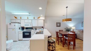 A compact kitchen with white cabinets, a microwave, a stove, and a sink. There is a dining area with a wooden table and chairs, and a hanging light fixture above. - Secluded Paradise Serene 2BR Golf Course Views (Pawleys Island)