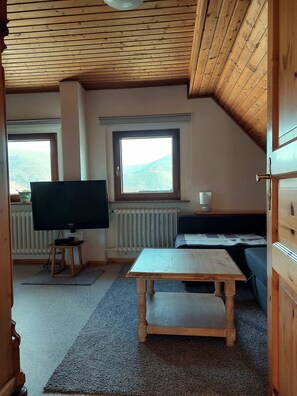 Ein gemütliches Zimmer mit Fernseher, kleinem Tisch, Sofa und Fenster mit Blick auf eine hügelige Landschaft.