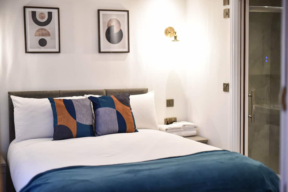 A neatly made bed with a blue blanket, two pillows, and a bedside table with a lamp and a folded towel.