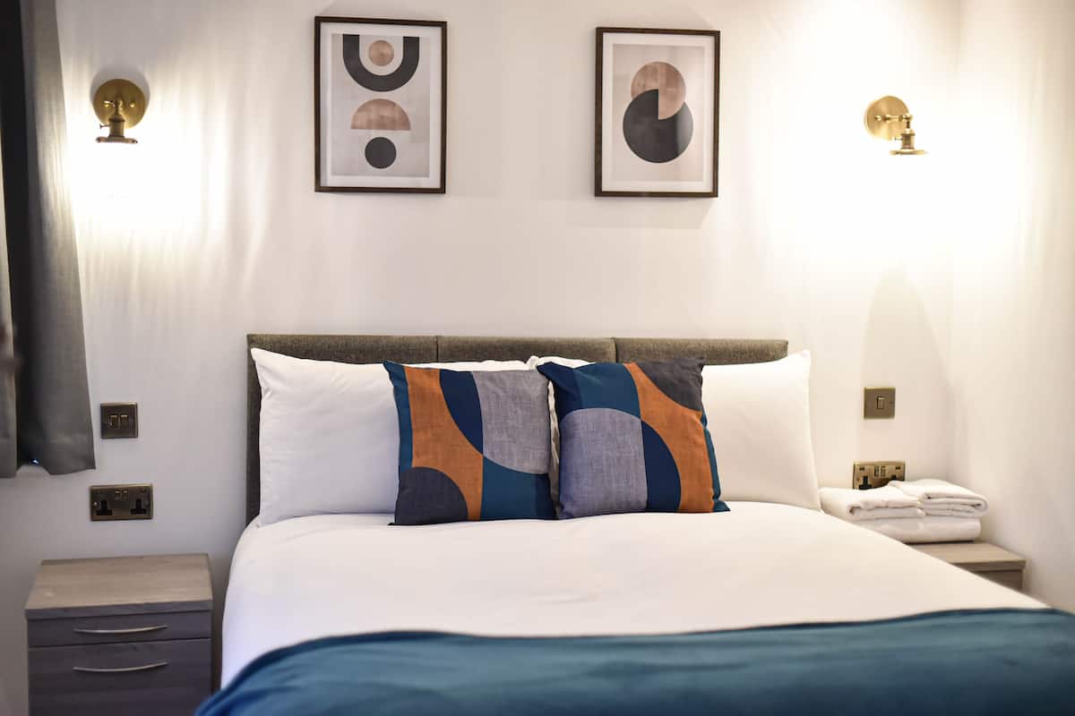 A neatly made bed with a white and blue bedspread, two decorative pillows, and a bedside table with a phone and a folded towel.
