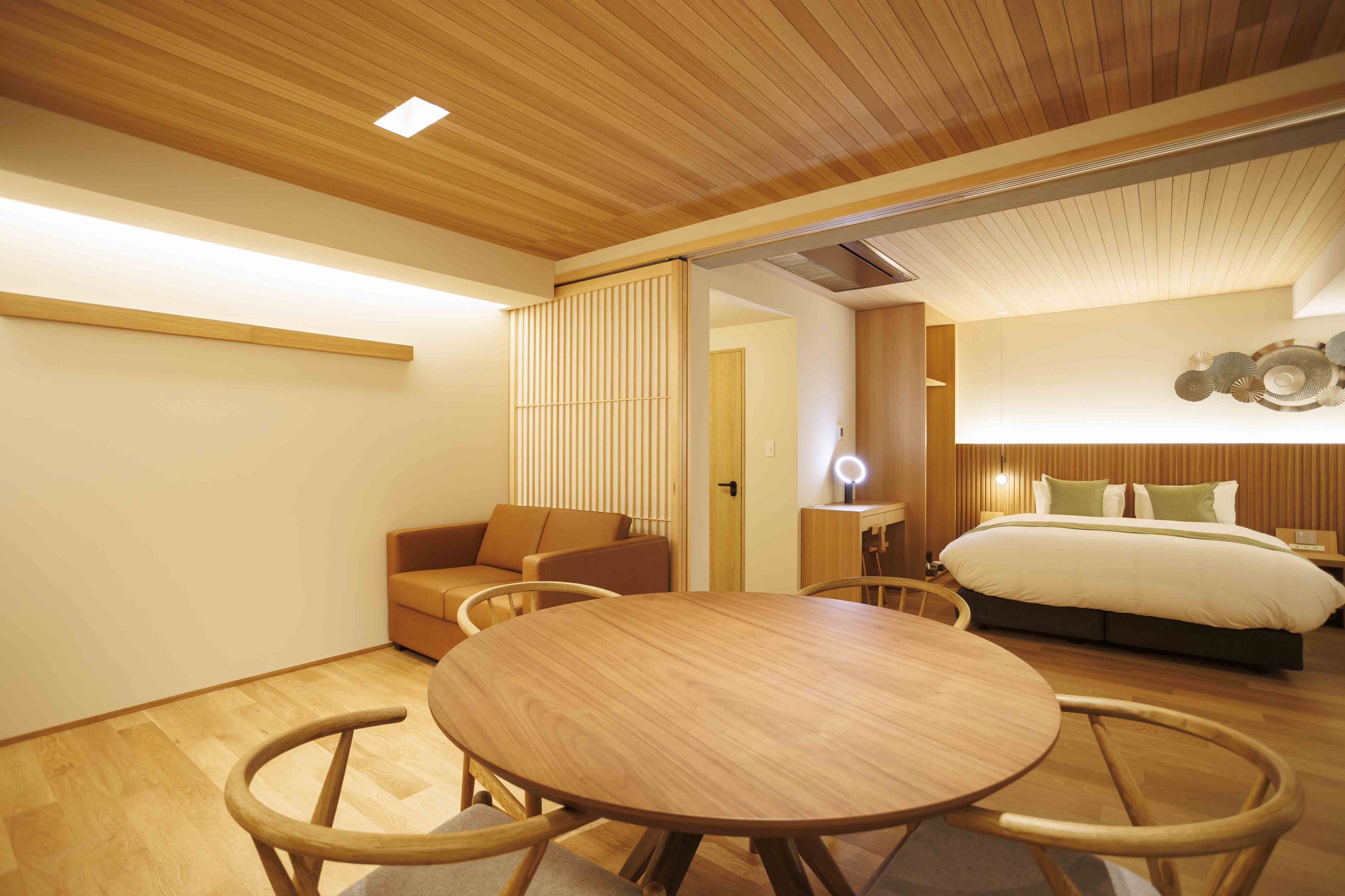 Une chambre d’hôtel moderne avec un plafond en bois, une table à manger ronde entourée de chaises, un canapé et un lit doté d’une tête de lit.