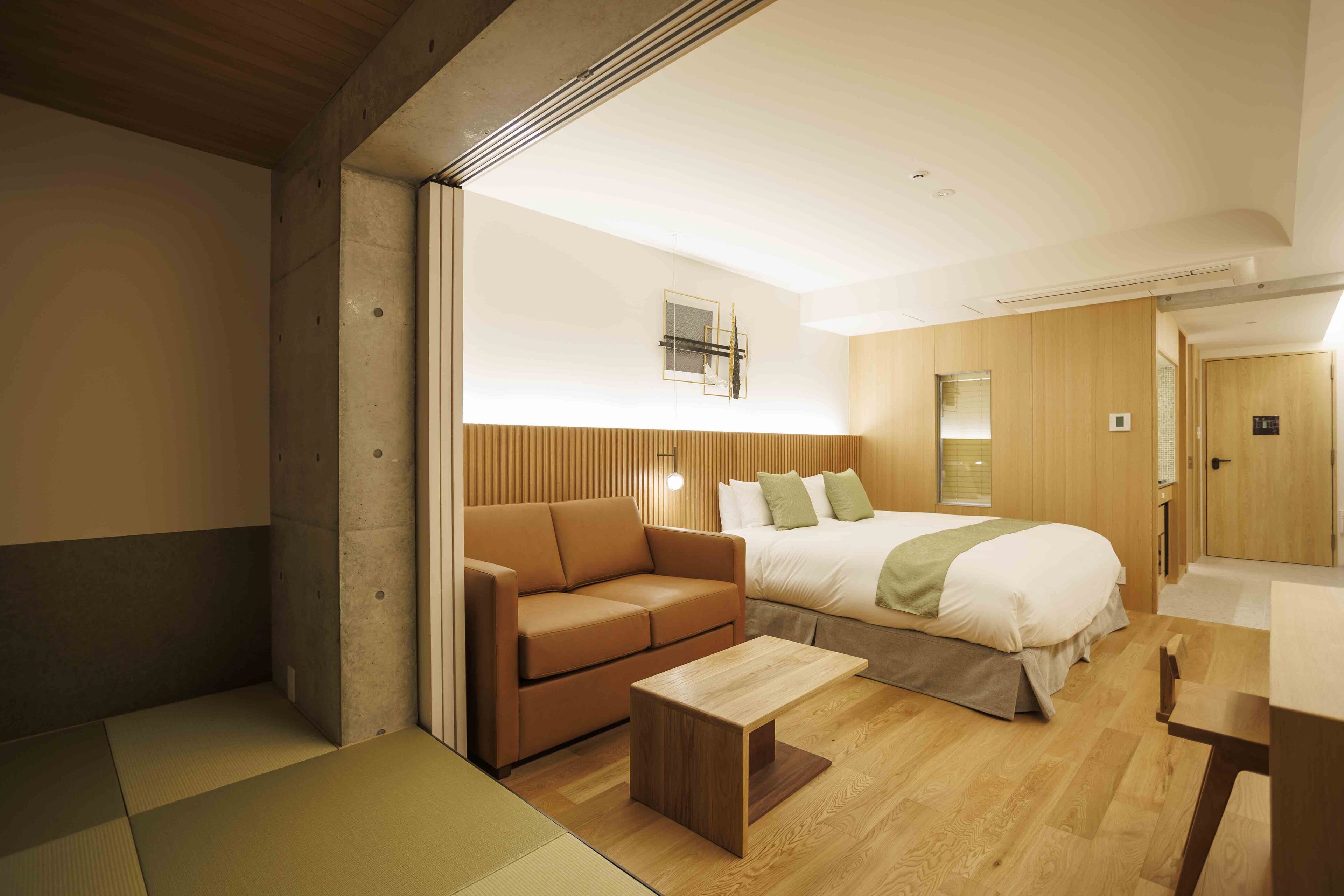 A modern hotel room with a wooden floor, a sofa, a bed with a green throw, and a small wooden table.