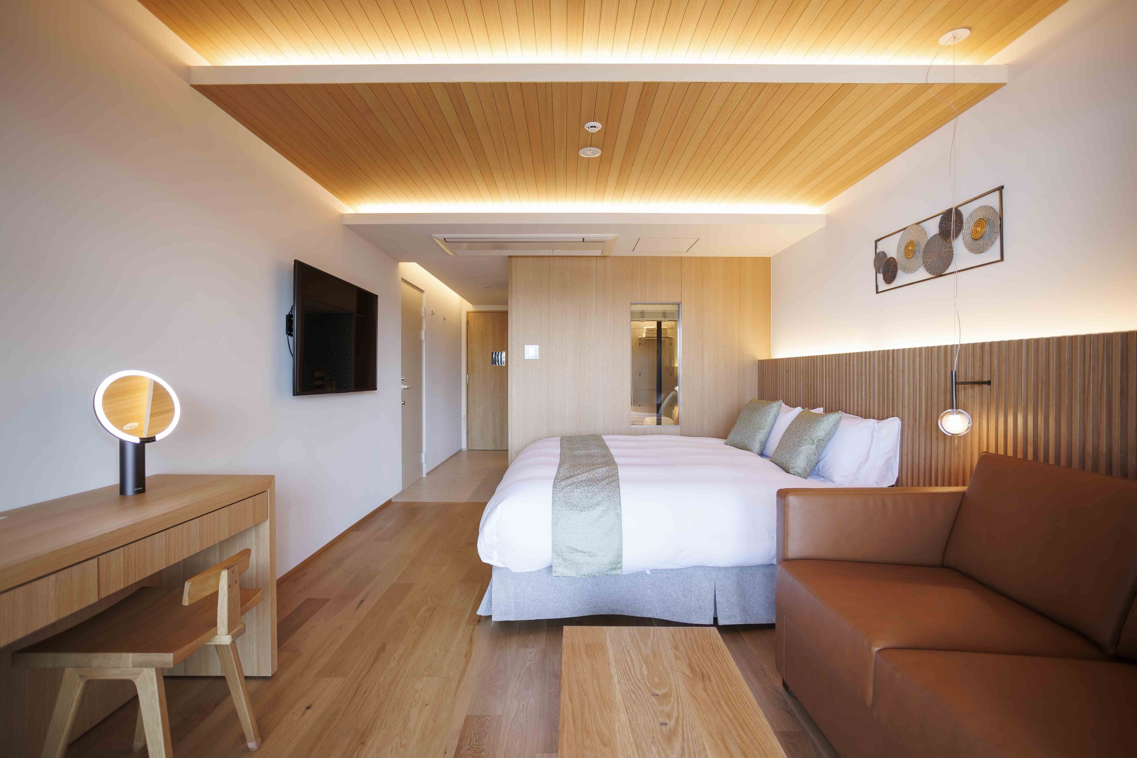 Ein modernes Hotelzimmer mit Holzboden, einem Bett mit grauer Bettdecke, einem braunen Ledersofa, einem Holztisch mit Stuhl und einem an der Wand befestigten Fernseher.