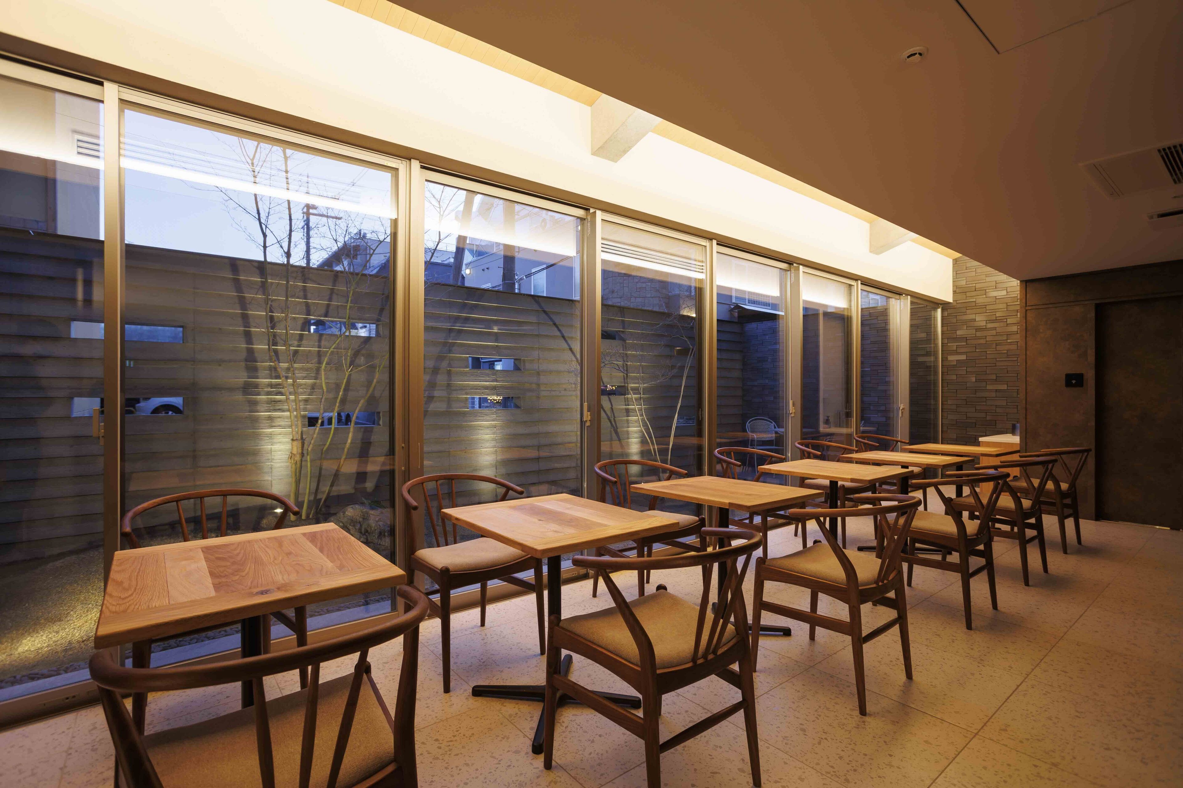 Un área de estar interior moderna con mesas y sillas de madera, ventanas amplias y vista a un edificio.