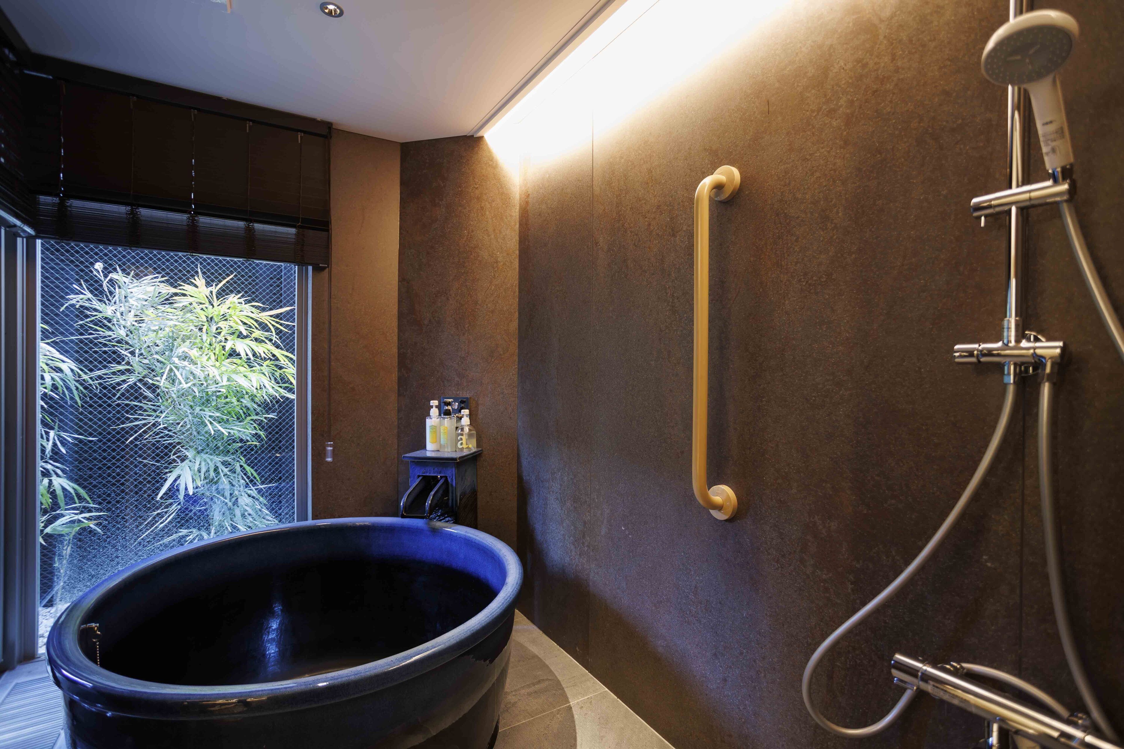 A modern bathroom with a large black bathtub, a handheld showerhead, and a glass door leading to a bamboo garden.