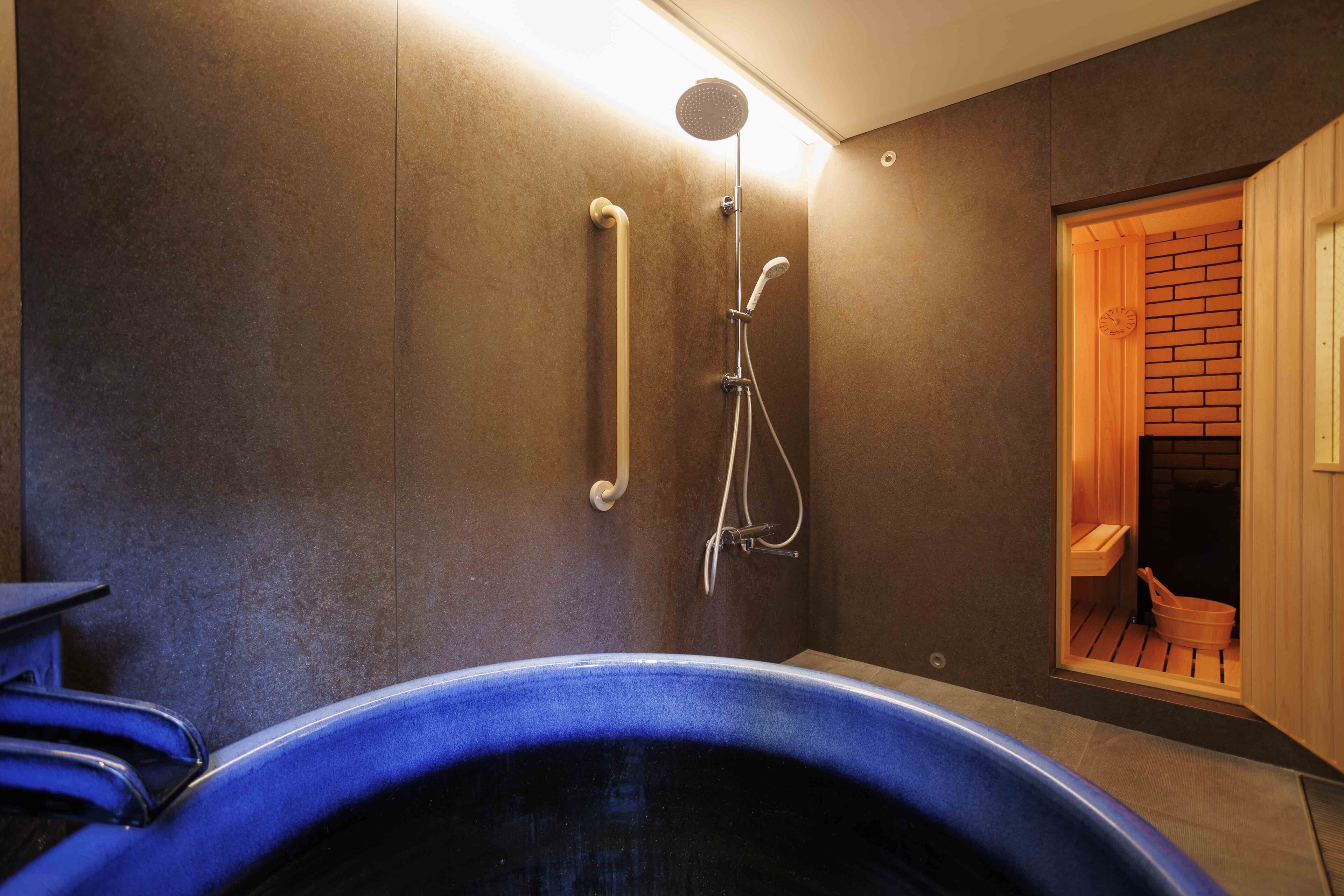 A modern bathroom with a large blue bathtub, a showerhead, and a wooden sauna door.