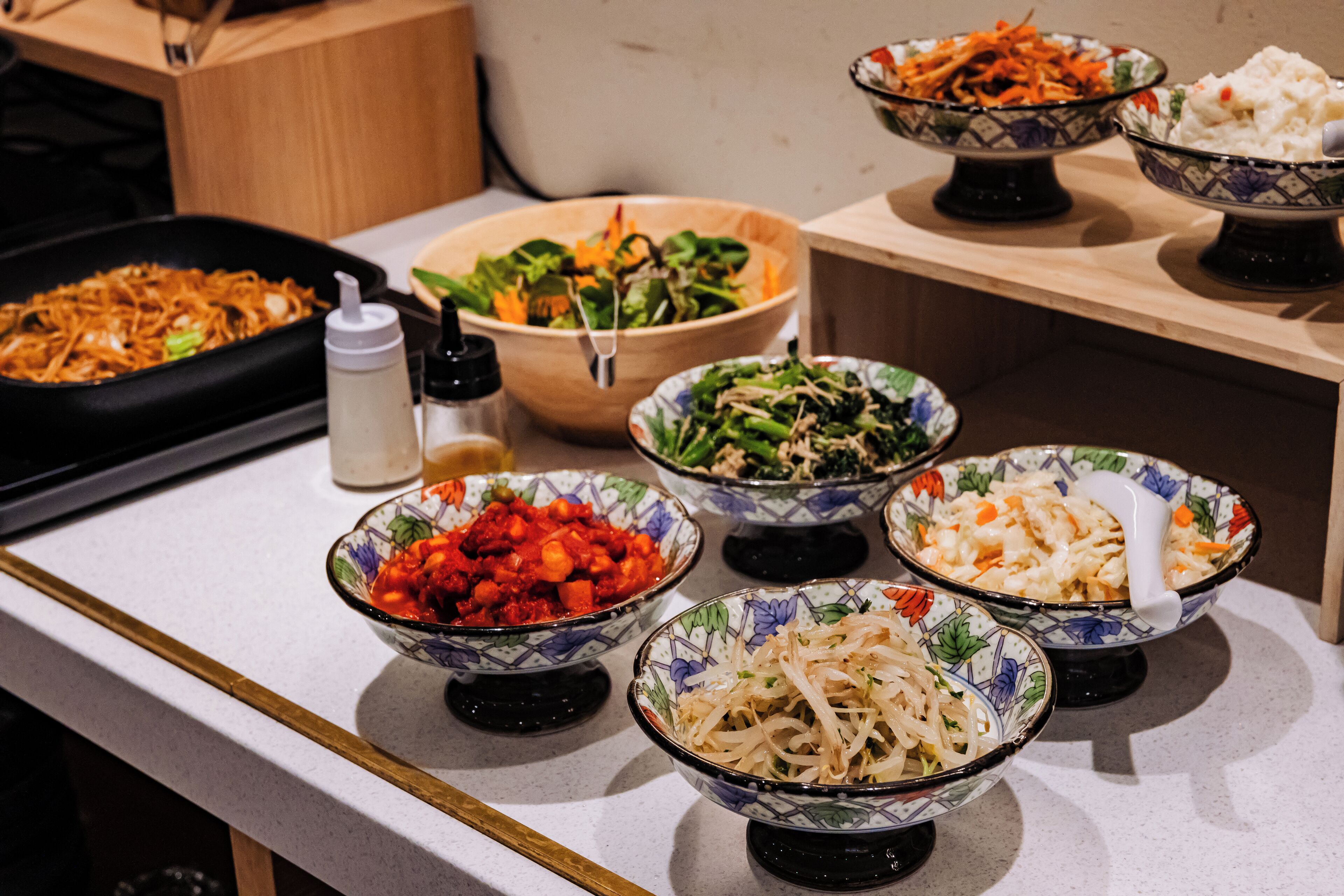 Une table de buffet proposant divers plats présentés dans des bols, notamment des salades, des nouilles et des légumes sautés.