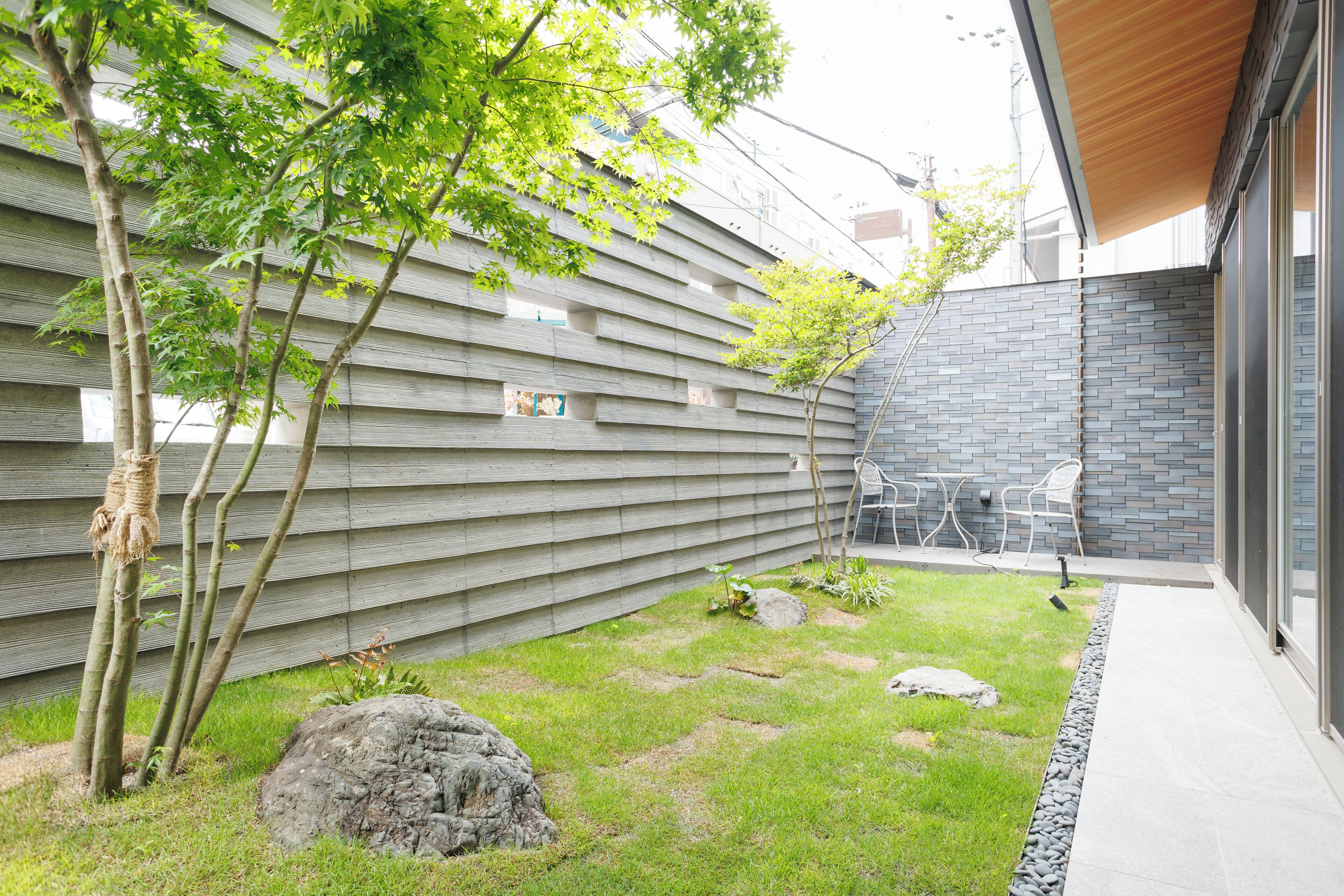 Un bâtiment moderne doté d’un mur à lattes de bois, d’une zone herbeuse parsemée de rochers et d’un petit jardin avec un arbre.