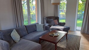 Un salon moderne avec un canapé gris, une table basse en bois et une vue sur un jardin grâce à de grandes fenêtres.