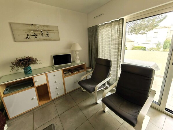 Ein Zimmer mit einem Glastisch, einem Computer, einer Lampe, zwei Stühlen, einem Gemälde und Blick durch ein Fenster.