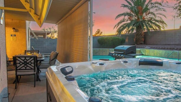 A hot tub with a patio area, a table with chairs, and a pool in the background. - 4BR Pool, Spa & Retro Arcade Polished Vegas Stay (North Las Vegas)