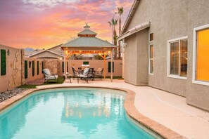 A swimming pool with a covered patio area, outdoor furniture, and a sunset view.