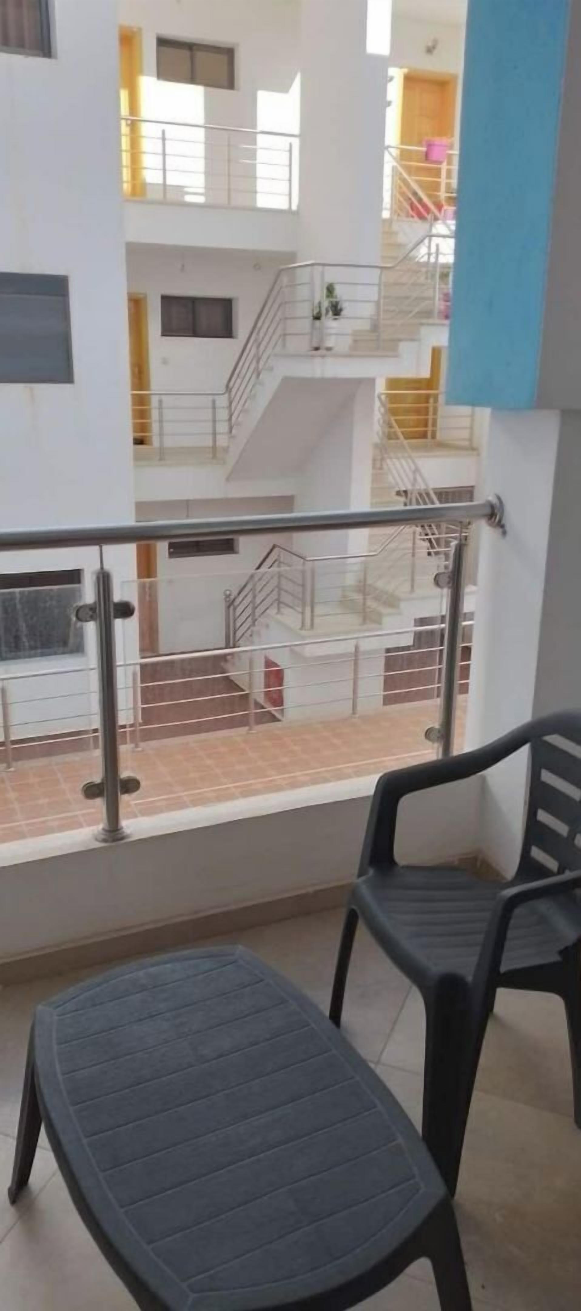 A balcony with a metal railing, two plastic chairs, and a view of a multi-story building with balconies and stairs.