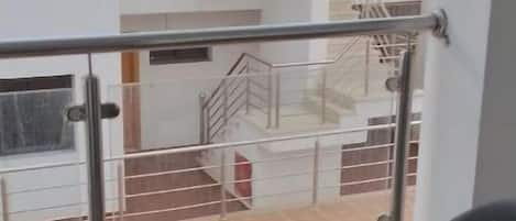 A balcony with a metal railing, two plastic chairs, and a view of a multi-story building with balconies and stairs.