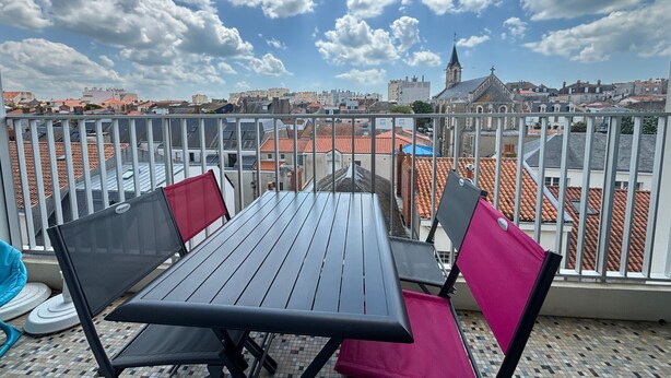 A balcony with a table and chairs, overlooking a cityscape with buildings and a church tower. - Renovated 2BR Apartment, Sleeps 5, City Center, Close to Beach, Parking, WiFi (Les Sables-d'Olonne)