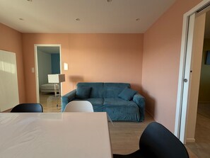 Een kamer met een bank, een tafel en stoelen. Een deur leidt naar een andere kamer.