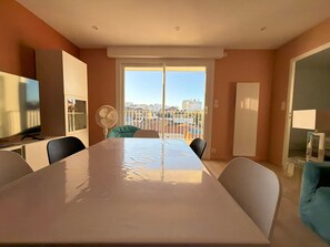 A dining area with a table, chairs, a fan, and a view of buildings through a large window. - Renovated 2BR Apartment, Sleeps 5, City Center, Close to Beach, Parking, WiFi (Les Sables-d'Olonne)