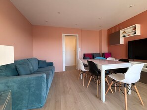 A modern living room with a blue sofa, a dining area with a white table and chairs, and a kitchen area with a TV and shelves. - Renovated 2BR Apartment, Sleeps 5, City Center, Close to Beach, Parking, WiFi (Les Sables-d'Olonne)