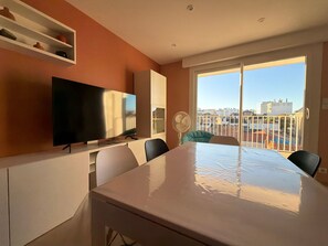 A modern living room with a flat-screen TV, a dining table, chairs, a white fan, and a large window with a city view. - Renovated 2BR Apartment, Sleeps 5, City Center, Close to Beach, Parking, WiFi (Les Sables-d'Olonne)