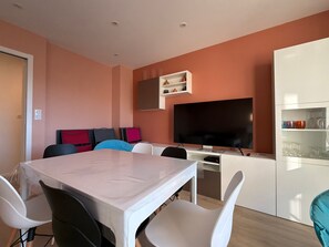 A modern living room with a dining table, chairs, a flat-screen TV, and a built-in cabinet with glassware. - Renovated 2BR Apartment, Sleeps 5, City Center, Close to Beach, Parking, WiFi (Les Sables-d'Olonne)