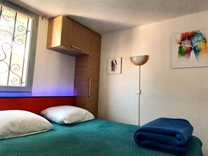 Um quarto com uma cama, almofadas, um guarda-roupa de madeira, uma janela com grades decorativas, um espelho fixado na parede e um quadro.