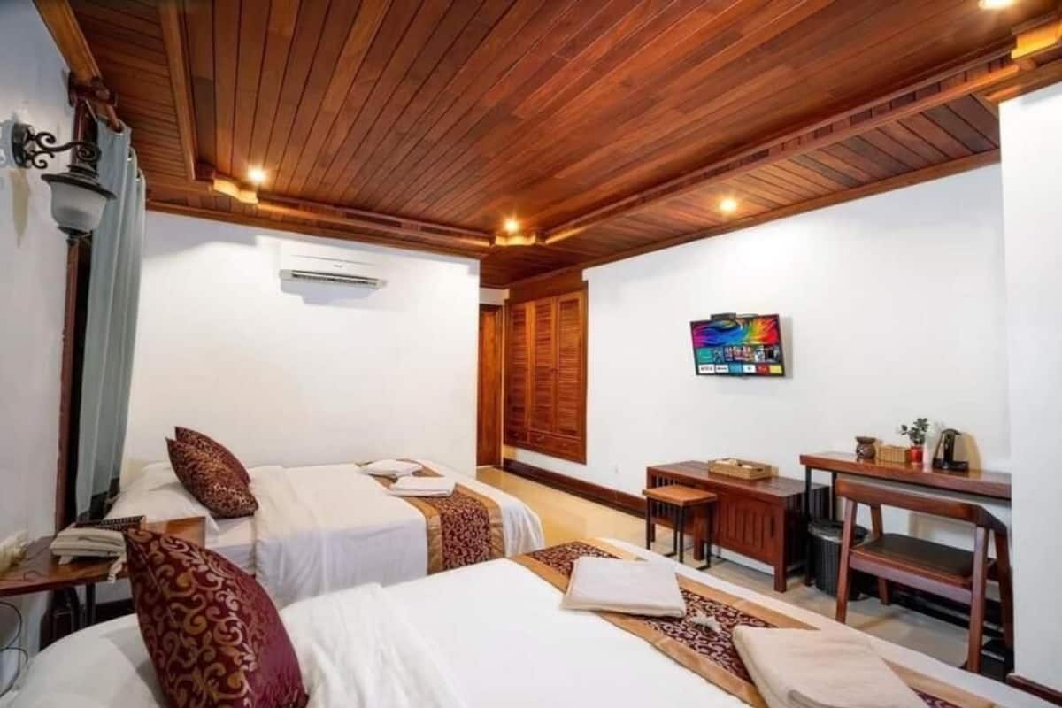 A room with two beds, wooden ceiling, a desk, and a wall-mounted TV.