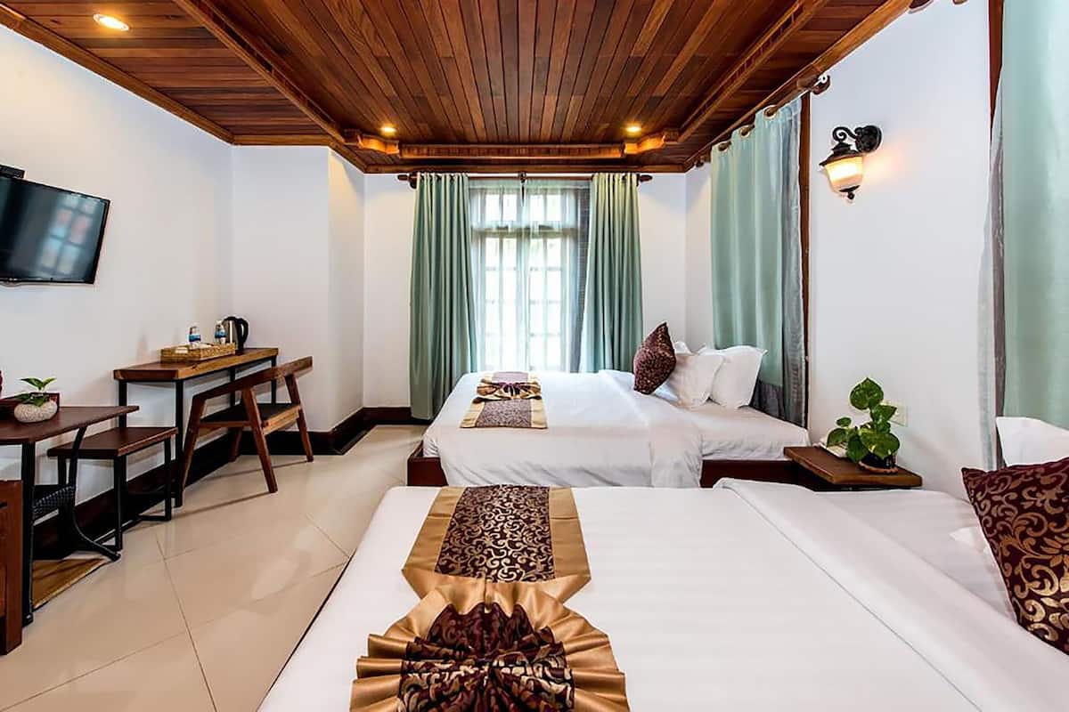 A hotel room with a wooden ceiling, a TV, a desk with a chair, two beds with pillows, and a plant.
