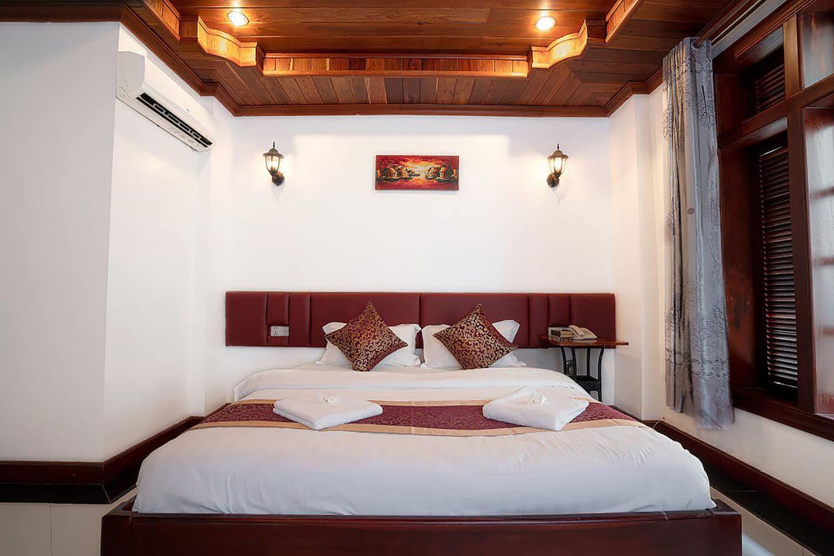 A hotel room with a large bed, wooden headboard, two wall-mounted lamps, a painting above the bed, and a window with curtains.