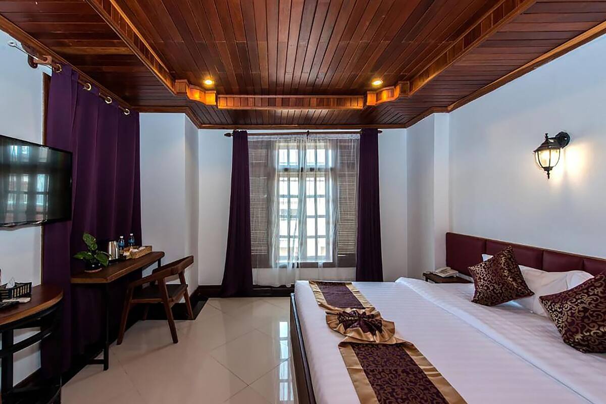 A hotel room with a wooden ceiling, a bed with a patterned bedspread, a desk with a chair, a television, and a window with curtains.
