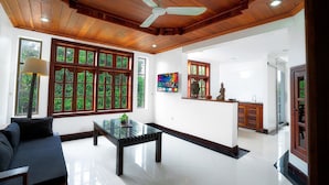 Amplia sala de estar con techo de madera, ventanas grandes, una mesa de centro de vidrio y una cocina visible a través de una puerta abierta.