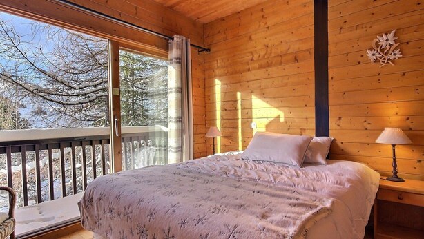 A cozy wooden cabin bedroom with a large bed, a nightstand, a lamp, and a window overlooking a snowy landscape. - Cozy chalet near slopes with Wifi (La Plagne)