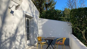 Eine kleine Terrasse mit gefliestem Boden, zwei gelbe Stühle, einem Holztisch und einer weißen Wand mit einer Tür und einem Fenster.