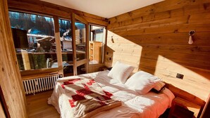 A wooden cabin bedroom with a large bed, a radiator, and a window overlooking a snowy landscape.