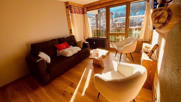 A living room with a dark brown sofa, a white armchair, and a wooden coffee table. There is a window with curtains and a view of buildings outside.