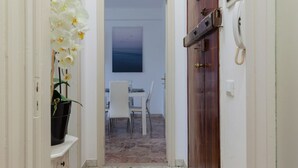 A narrow hallway with a potted orchid, a dining area with chairs and a table, and a wooden door with a telephone.