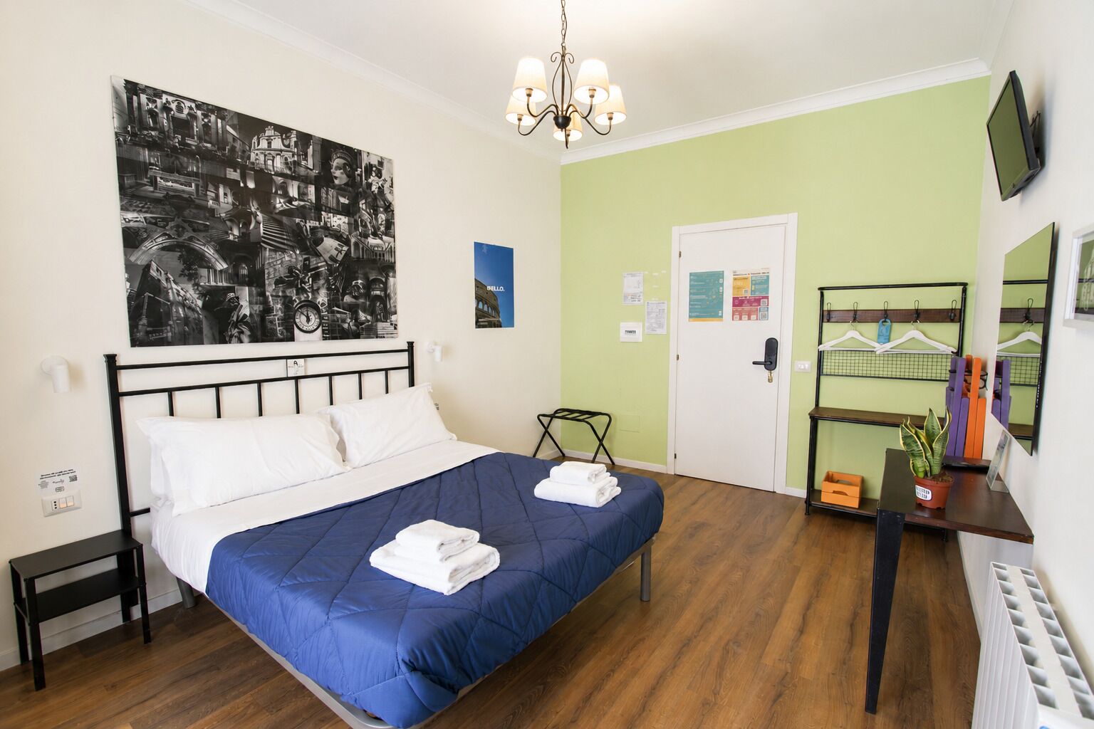 Une chambre à coucher avec un lit, un téléviseur fixé au mur, une plante suspendue et une porte ornée d’une affiche.