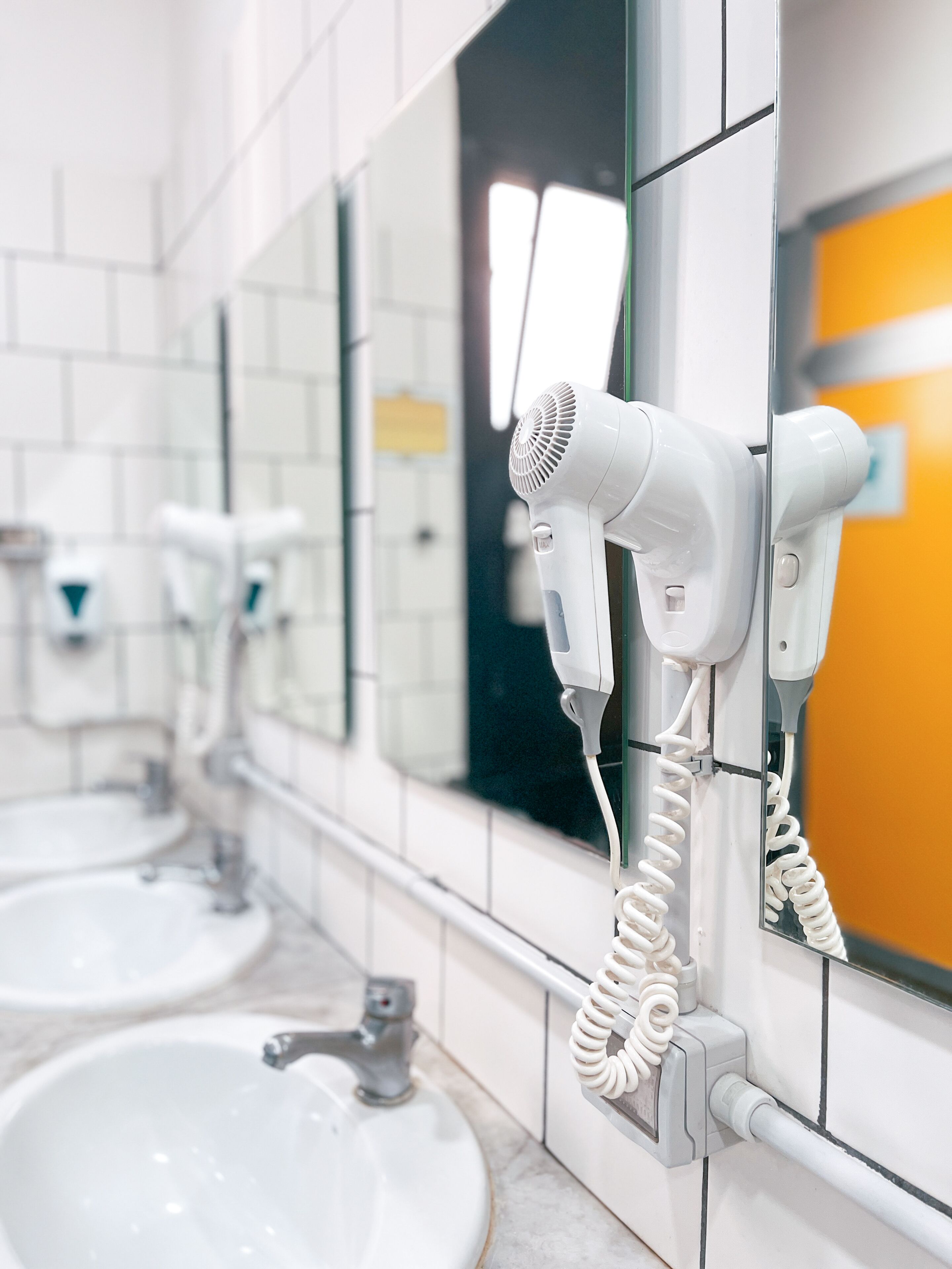 Un baño con paredes de azulejos blancos, dos lavamanos y un secador de pelo fijado a la pared.