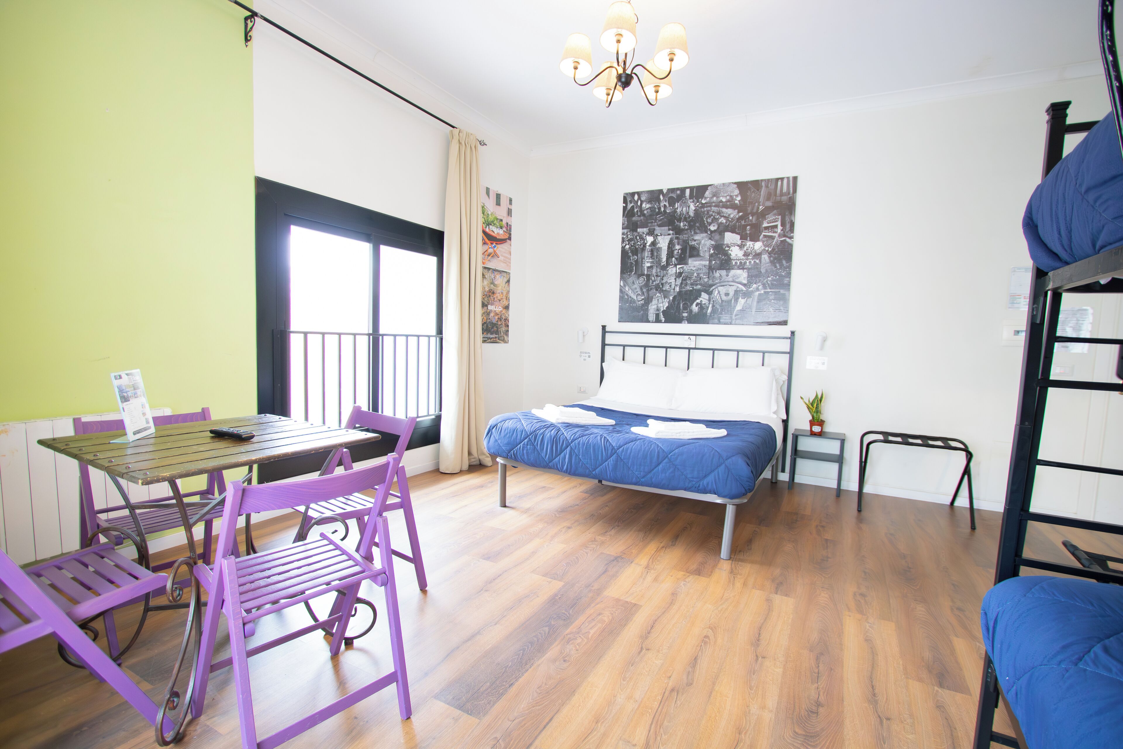 Una habitación con una cama, una mesa con sillas, una mesita auxiliar pequeña, una lámpara colgante y un cartel grande en la pared.