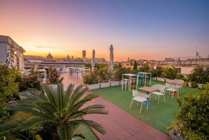 Un patio al aire libre con mesas y sillas, rodeado de follaje, con vistas a un paisaje urbano que incluye edificios y un río al atardecer.