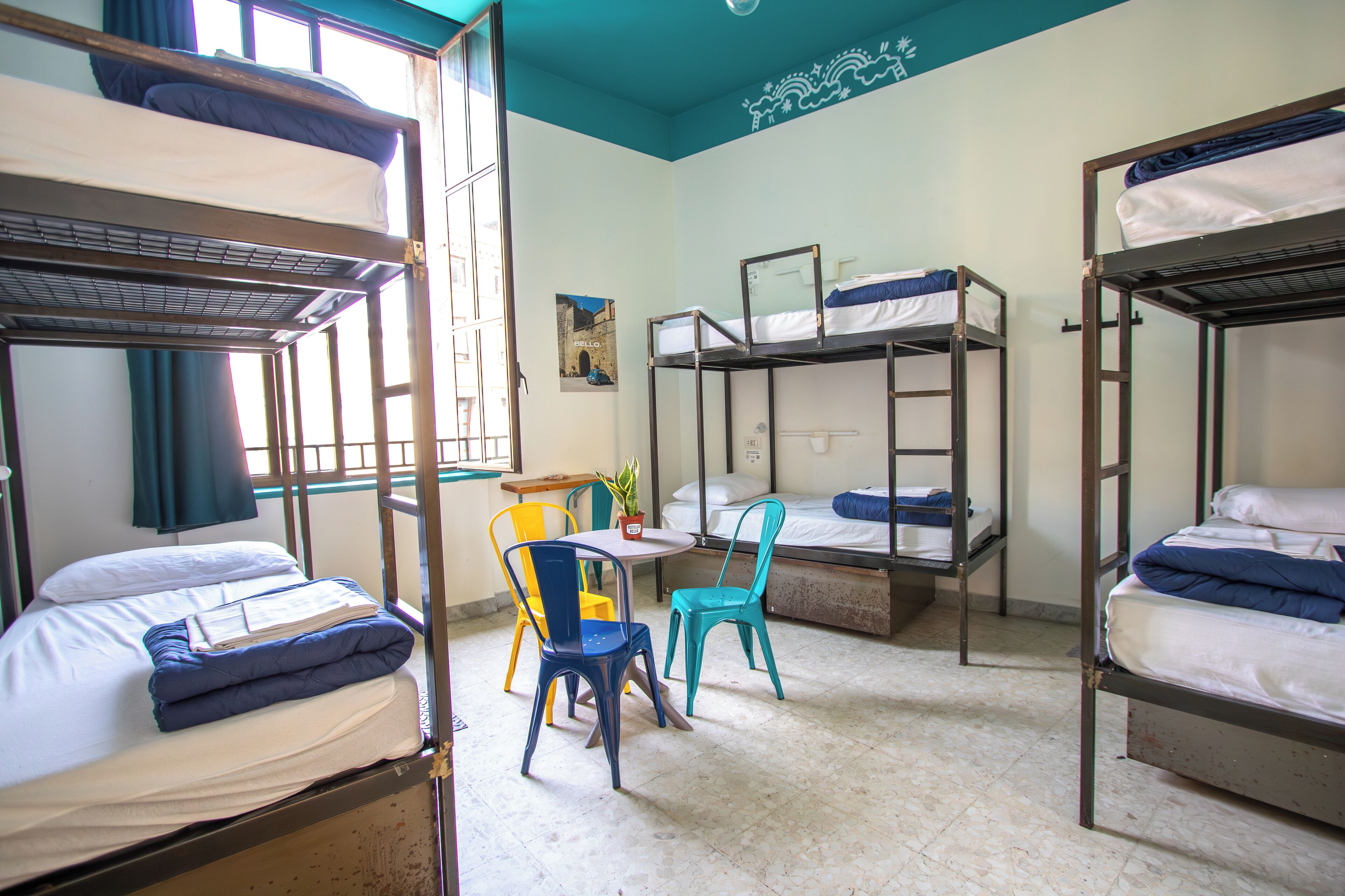 A dormitory room with bunk beds, a small table, chairs, and a window with curtains.
