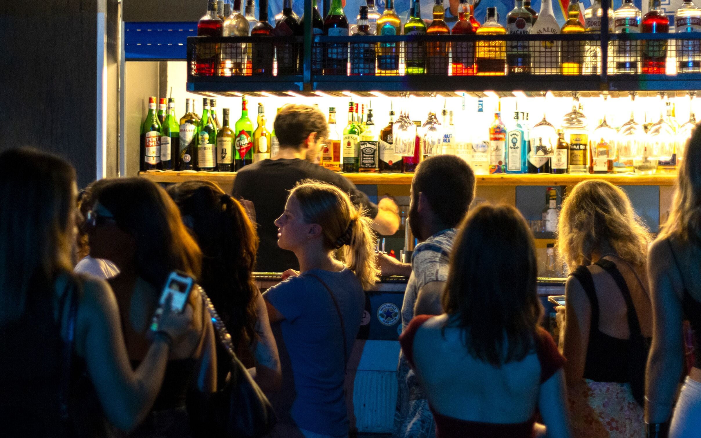 A bar area with various bottles and glasses, and people standing around.