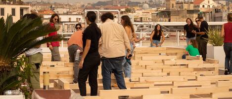 A group of people on a rooftop with wooden seating areas and a cityscape in the background.